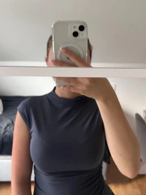 Mörkgrå ärmlös topp med hög hals - Snygg mörkgrå topp med hög hals och ärmlös design. Toppen har en tight passform och är gjord i ett mjukt, stretchigt material som sitter skönt mot kroppen. Perfekt att styla med jeans eller kjol för en clean look.