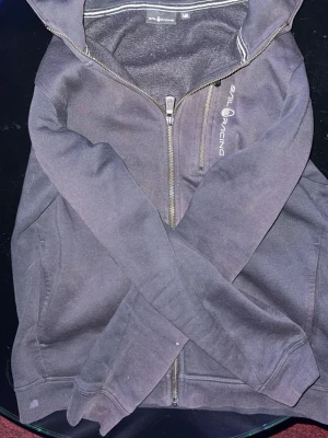 Mörkgrå hoodie från Sail Racing - Mörkgrå hoodie från Sail Racing med dragkedja framtill och en extra bröstficka med dragkedja. Snygg logga på bröstet och mjukt, borstat innerfoder. Klassisk huva och ribbade muddar vid ärmar och nederkant. Perfekt för en avslappnad och sportig stil.