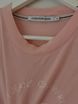 Rosa Calvin Klein Jeans t-shirt - Snygg rosa t-shirt från Calvin Klein Jeans med vit logga och text framtill. Klassisk rund halsringning och korta ärmar. Tillverkad i mjuk bomull som känns skön mot huden. Perfekt för en avslappnad och trendig look.