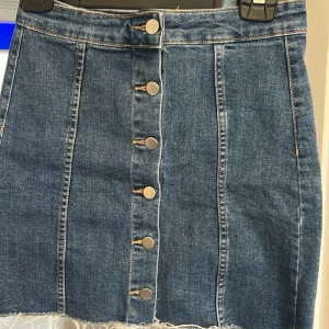 Blå jeanskjol med knappar framtill - Snygg blå jeanskjol i kort modell med rå kant nertill och dekorativa sömmar. Kjolen har knappar hela vägen framtill💕