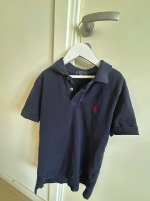 Marinblå pikétopp från Polo Ralph Lauren - Klassisk mörkblå pikétröja från Polo Ralph Lauren med korta ärmar och den ikoniska röda logotypen broderad på bröstet. Tröjan har krage och två knappar.