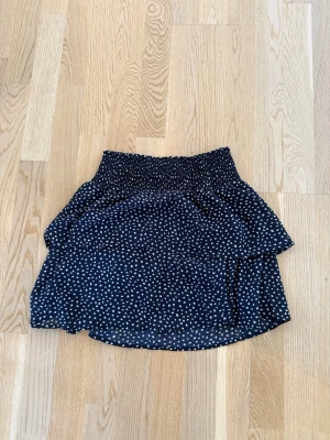 Svart prickig kjol från ONLY - Kort svart kjol från ONLY med vita prickar och volangdetaljer. Kjolen har bred resår i midjan för skön passform och är tillverkad i ett lätt, luftigt material som passar perfekt till varmare dagar. Kan sänka pris vid snabb affär.