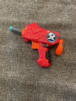 Röd leksakspistol Zuru X-Shot - Rolig röd leksakspistol från Zuru X-Shot med svart och orange detaljer samt blått märke på sidan. Pistolen är gjord i plast och har en futuristisk design. Perfekt för lek och äventyr med kompisar.