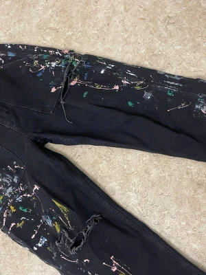 Svarta jeans med färgstänk och slitningar - Unika svarta jeans med färgstänk i rosa, grönt, gult och vitt. Jeansen har slitna detaljer vid knäna och en avslappnad passform. Perfekta för dig som vill sticka ut med en kreativ och edgy stil. Materialet är klassisk denim.