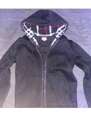 Svart hoodie från Burberry med dragkedja - Säljer en svart hoodie från Burberry med dragkedja och klassiskt rutigt foder i huvan. Hoodien har fickor framtill och justerbara snören i huvan. Perfekt för dig som gillar stilrena plagg med exklusiv känsla och snygga detaljer.