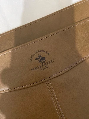 Brun datorväska Santa Barbara Polo - Stilren brun datorväska i konstläder från Santa Barbara Polo & Racquet Club. Perfekt för att bära din laptop på ett snyggt och organiserat sätt.