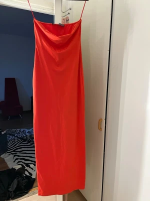 Orange långklänning med tunna band - Säljer en snygg orange långklänning med tunna axelband och draperad urringning. Klänningen har en rak passform och är perfekt för sommarens alla tillfällen. Materialet känns mjukt och har ett lätt glansigt utseende.