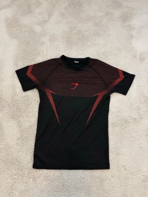 Svart och röd T-shirt  - Snygg svart träningströja med röda grafiska detaljer på bröst, axlar och rygg. Tillverkad i ett stretchigt och snabbtorkande material som passar perfekt för gymmet eller löpning. Kortärmad modell med rund hals och tight passform.