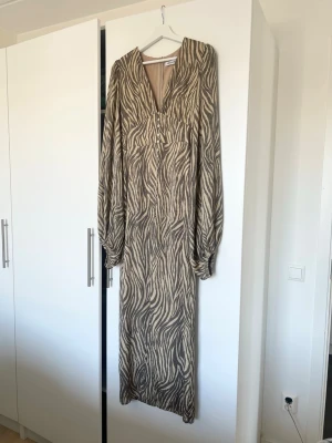 Zebramönstrad långklänning från Adoore - Långklänning från Adoore med zebramönster i beige och brunt. Klänningen har v-ringning, knappar framtill och långa, lösa ärmar. Tillverkad i ett mjukt, lätt material som ger ett snyggt fall. Perfekt för dig som gillar statementplagg. Använd endast vid ett tillfälle. 