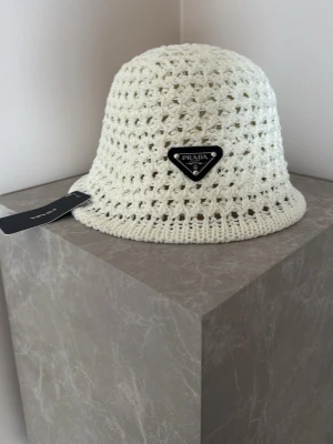 Vit virkad bucket hat från Prada - Snygg vit bucket hat från Prada i virkat material med ett ikoniskt svart logomärke framtill. Hatten har en luftig och trendig design som passar perfekt för att lyfta din stil. Perfekt accessoar för dig som gillar exklusiva och unika detaljer.