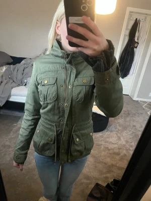 Grön parkajacka med fickor - Snygg grön parkajacka med fyra framfickor och dragkedja samt knappar. Jackan har hög krage, justerbar midja med snörning och guldiga detaljer. Perfekt för lager på lager och en avslappnad streetstil. De går att sänka pris 