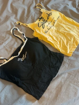 Två ribbade linnetoppar med spets - Säljer två snygga linnetoppar, en svart med vit spetskant och broderad text, samt en gul med tryckt text och liten rosett fram. Båda har smala axelband och är perfekta för sommaren. Materialet känns mjukt och stretchigt, troligen bomull eller polyester.