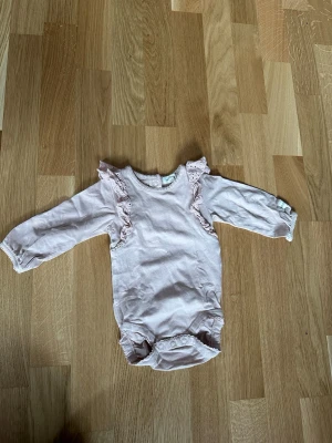 Ljusrosa body med volangdetaljer - Supersöt ljusrosa body med långa ärmar och volangdetaljer längs axlarna. Knäppning finns både i grenen och bak i nacken för enkel påklädning. Tillverkad i mjukt tyg som känns skönt mot huden. Perfekt för dig som gillar gulliga och feminina detaljer.