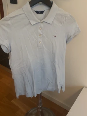 Ljusblå pikétröja från Gant - Klassisk ljusblå pikétröja från Gant med korta ärmar, krage och knappar framtill. Liten broderad logga på bröstet. Tillverkad i mjuk bomull som känns skön mot huden. Perfekt till jeans eller kjol för en avslappnad look.