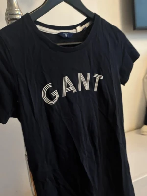 Marinblå GANT t-shirt med tryck - Snygg marinblå t-shirt från GANT med vit logotyp framtill. Klassisk rund halsringning och korta ärmar. Tillverkad i mjuk bomull som känns skön mot huden. Perfekt till jeans eller shorts för en avslappnad look.