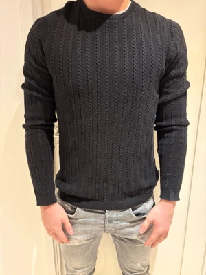 Svart kabelstickad tröja herr - Snygg svart kabelstickad tröja med rund hals och långa ärmar. Tröjan har ett klassiskt mönster med vertikala flätor och ribbade muddar vid ärmslut och nederkant. Perfekt för dig som gillar stilrena och tidlösa plagg.
