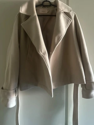 Beige kappa från Nelly med skärp - Stilren beige kappa från Nelly med bred krage, långa ärmar och tillhörande skärp i midjan. Kappan har fickor på sidorna och en mjuk, ull-liknande yta som ger en lyxig känsla. Perfekt för att höja din outfit under kyliga dagar.
