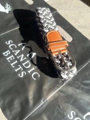 Svartvitt flätat bälte Scandic Belts - Snyggt svartvitt flätat bälte med detaljer i brunt skinn och silverfärgat spänne. Perfekt accessoar för att lyfta din outfit med en modern touch. Bältet har ett randigt mönster och klassisk design. 105cm