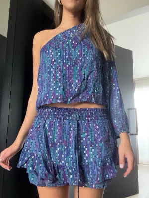 Set från Brownie - Säljer ett färgglatt set från Brownie med blå, lila och turkosa toner samt silverdetaljer. Toppen har en axel och lång ärm, kjolen är kort med volanger och resår i midjan. Materialet är lätt och luftigt, perfekt för en trendig look. Helt i ny skick 