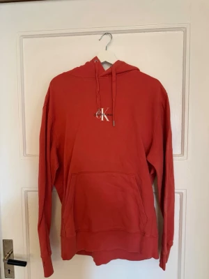 Röd hoodie från Calvin Klein - Snygg röd/orange hoodie från Calvin Klein med klassisk logga på bröstet. Hoodien har huva med dragsko och en stor magficka framtill. Tillverkad i mjukt bomullsmaterial och har ribbade muddar vid ärmslut och nederkant. Perfekt för en avslappnad och trendig look.