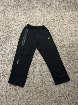 Svarta NIKE NOCTA byxor - Säljer ett par svarta träningsbyxor från NIKE NOCTA.  gul logga och detaljer. Byxorna har resår i midjan och raka ben. Perfekta för träning eller chill. Materialet känns lätt och bekvämt, och det finns en diskret ficka på ena benet. Tveka inte på att höra av dig vid minsta fundering!