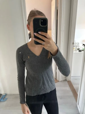 Grå stickad tröja från Ralph Lauren - Grå stickad tröja från Ralph Lauren med v-ringning och lång ärm. Tröjan har en liten broderad logga på bröstet. (Äkta)
