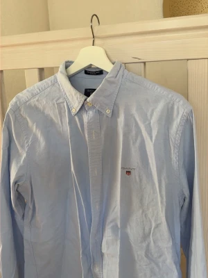Ljusblå skjorta från Gant - Klassisk ljusblå skjorta från Gant med button down-krage och broderad logga på bröstet. Skjortan har lång ärm, vita knappar och är tillverkad i mjuk bomull. Perfekt för en clean och stilren look. Storlek L men passar M lika bra! 