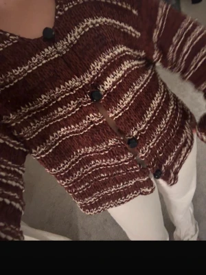 Randig vinröd stickad kofta - Säljer en stickad kofta i vinrött med beige och vita ränder. Koftan har rund halsringning, stora svarta knappar och lång ärm. Perfekt för lager på lager och riktigt mysig. Materialet är grovstickat och ger en avslappnad vibe.