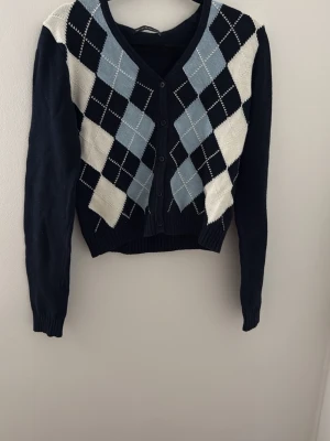 Kofta med argylemönster i blått och vitt - Snygg kofta med klassiskt argylemönster i svart, ljusblått och vitt. Koftan har V-ringning, knappar framtill och ribbade muddar. Croppad modell som passar perfekt till high waist-jeans. Materialet känns mjukt och stretchigt.