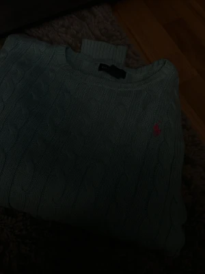 Ljusblå kabelstickad tröja Ralph Lauren - Snygg ljusblå kabelstickad tröja från Ralph Lauren med rund halsringning och klassisk rosa logga broderad på bröstet. Tröjan har ribbade muddar och är perfekt för dig som gillar preppy stil och vill ha något både bekvämt och stilrent.