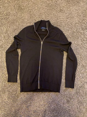 Mörkblå merinoullströja från Riley - Snygg svart tröja från Riley i mjuk merinoull. Tröjan har hel dragkedja framtill, hög krage och ribbade muddar vid ärmslut och nederkant. Perfekt för lager på lager och stilrena outfits.