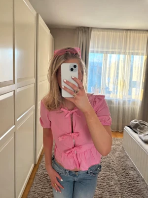 Rosa topp med rosetter och volang - Supersöt rosa topp med korta ärmar, tre dekorativa rosetter framtill och volangkant nedtill. Toppen har en croppad passform och är tillverkad i ett lätt bomullstyg. Perfekt för dig som vill ha en gullig och trendig look.