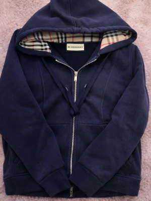 Mörkblå hoodie från Burberry - Snygg mörkblå hoodie från Burberry med dragkedja och klassiskt rutigt foder i huvan. Tillverkad i mjukt material och har ribbade muddar vid ärmar och nederkant. Perfekt för dig som vill ha en stilren och exklusiv look.