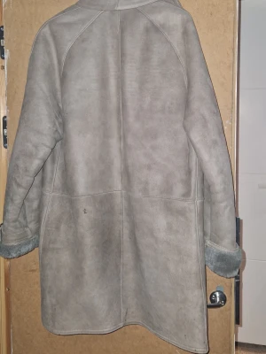 Grå äkta fårskinnskappa med uppvikta ärmar - Snygg grå fårskinnskappa med mjukt foder och uppvikta ärmslut. Kappan har två stora fickor framtill och stängs med knappar. Perfekt för kalla dagar och ger en klassisk look med sin raka passform och diskreta sömmar.