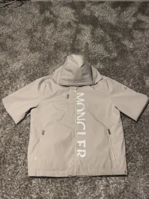ljus grå vindjacka från Moncler - Snygg ljus grå vindjacka från Moncler med stor logga framtill, dragkedja och två fickor med dragkedja. Jackan har huva och justerbar nederkant med dragsko. Tillverkad i lätt och vattenavvisande material, perfekt för blåsiga dagar. ( Säljer billigare pågrund av blod fläck)