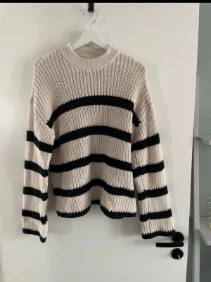 Beige stickad tröja med svarta ränder - Säljer en beige stickad tröja från Vero Moda med breda svarta ränder. Tröjan har rund halsringning, lång ärm och grovstickad struktur. Finns på fler sidor