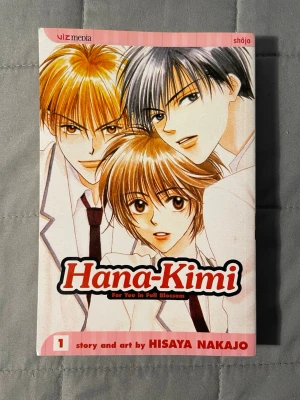 Hana-Kimi Volym 1 av Hisaya Nakajo - Manga-bok 'Hana-Kimi' volym 1 av Hisaya Nakajo. Omslaget har en ljusrosa bakgrund med illustration av tre ungdomar i skoluniform. Boken är på engelska och tillhör genren shōjo. Perfekt för dig som gillar romantik och drama i skolmiljö.