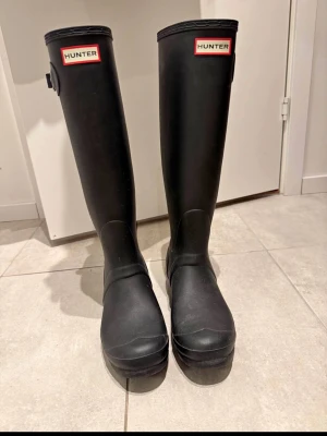 Svarta höga gummistövlar Hunter - Säljer ett par klassiska svarta höga gummistövlar från Hunter. De har en stilren design med logotyp framtill och justerbart spänne på sidan. Tillverkade i slitstarkt gummi som håller dig torr under regniga dagar.
