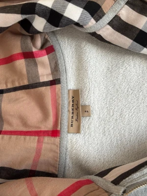 Grå hoodie från Burberry med dragkedja - Snygg grå hoodie från Burberry med klassiskt rutigt foder i huvan. Hoodien har dragkedja framtill, två fickor och justerbar huva med snören. Tillverkad i mjukt material som känns skönt mot huden. Perfekt för dig som vill ha en stilren och exklusiv look.