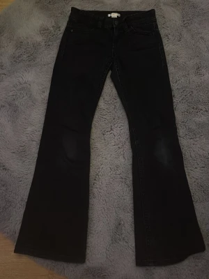 Svarta bootcut jeans med fickdetaljer - Snygga svarta bootcut jeans med klassiska fem fickor och coola ficklock bak med knappar. Jeansen har låg midja och utsvängda ben, perfekta för en trendig look. Materialet är denim med lite stretch för extra komfort.