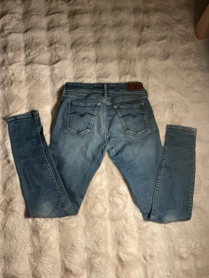 Ljusblå skinny jeans från Replay - Säljer ett par ljusblå skinny jeans från Replay med klassiska bakfickor och snygga slitningar. Jeansen har en läderpatch med Replay Blue Jeans-loggan bak i midjan och är tillverkade i mjukt denimtyg med stretch. Perfekta för en avslappnad och trendig look. W27 L30