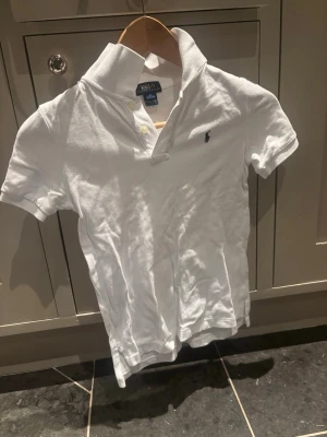Vit pikétröja från Ralph Lauren - Klassisk vit pikétröja från Ralph Lauren med korta ärmar och krage. Tröjan har två knappar vid halsen och den ikoniska lilla logotypen broderad på bröstet. Perfekt basic till garderoben.