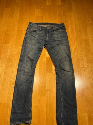 Nudie Jeans Tube Tom - Säljer ett par mörkblå jeans med raka ben och snygga slitningar på både fram- och baksida. Jeansen har klassisk femficksdesign, kontrastsömmar och är tillverkade i denim. Perfekta för en avslappnad och trendig stil.