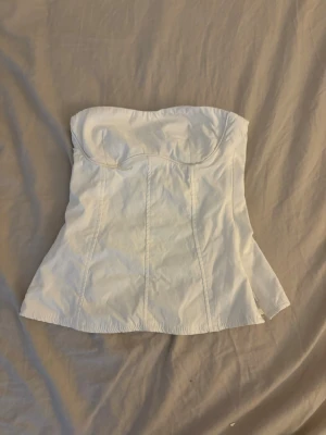 Vit axelbandslös korsettopp - Säljer en vit axelbandslös korsettopp med markerade sömmar och lätt utsvängd nederkant. Toppen har en figurnära passform och bygel vid bysten för extra stöd. Perfekt att styla med jeans eller kjol för en trendig look.