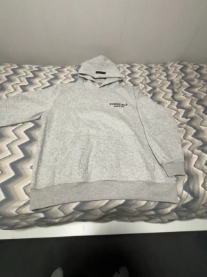 Grå hoodie Essentials Fear of God - Snygg grå hoodie från Essentials Fear of God med svart logga på bröstet. Klassisk passform med huva och känguruficka framtill. Tillverkad i mjukt bomullsmaterial som känns skönt mot huden. Perfekt för en avslappnad och trendig stil.