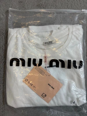 Vit  t-shirt med logga - Snygg vit t-shirt  med stor svart logga framtill. Klassisk rund hals och korta ärmar. Tillverkad i mjuk bomull för en skön känsla. Perfekt till jeans eller kjol för en clean och trendig look.