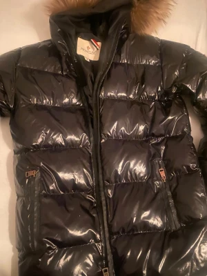 Svart glansig dunjacka från Moncler - Snygg svart dunjacka från Moncler med glansigt yttertyg och stor huva med fuskpäls. Jackan har två framfickor med dragkedja och Moncler-logga på insidan. Perfekt för kalla vinterdagar och riktigt trendig look.