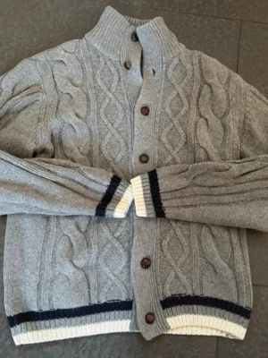 Grå kabelstickad kofta / cardigan från McNeal - Säljer en grå kabelstickad kofta från McNeal med bruna knappar och ribbad hög krage. Koftan har vita och svarta ränder vid ärmslut och nederkant, samt ett klassiskt mönster framtill. Perfekt för dig som gillar en stilren och mysig look.