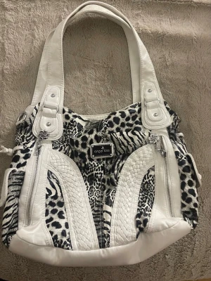 Vit och svart handväska Angel Kiss - Snygg handväska från Angel Kiss i vitt och svart med djurmönster. Väskan har flätade detaljer, flera dragkedjefickor och breda axelband. Materialet är konstläder och textil, perfekt för dig som gillar att sticka ut med din stil.