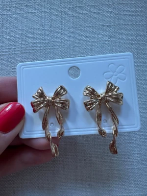 Guldiga rosettörhängen - Snygga örhängen i form av rosetter i guldfärgad metall. De har en tvinnad design på banden som ger en cool och trendig look. Perfekta för att lyfta vilken outfit som helst med en söt och lekfull touch.